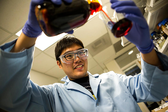 A man in a labcoat and goggles pouring dark liquid into a beaker.