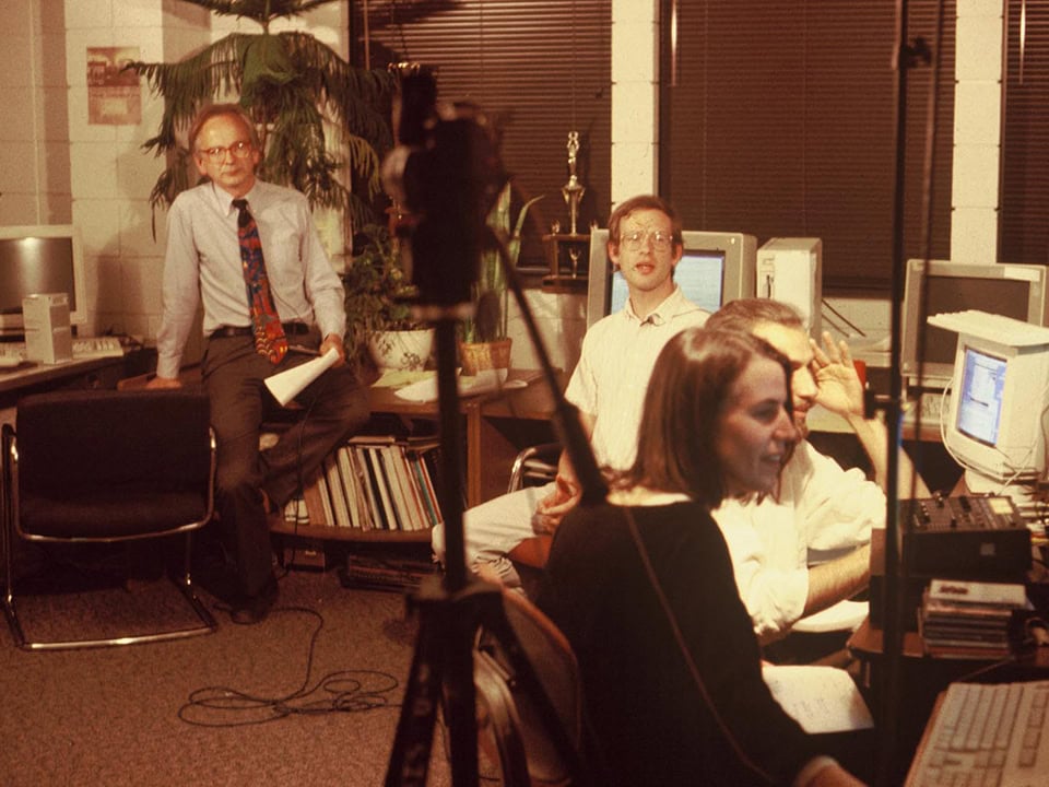 Orange-tinted old photo with Perry Samson, Jeff Masters and two other students in a room with 90's desktop computers and a camcorder on a tripod pointed at Perry.