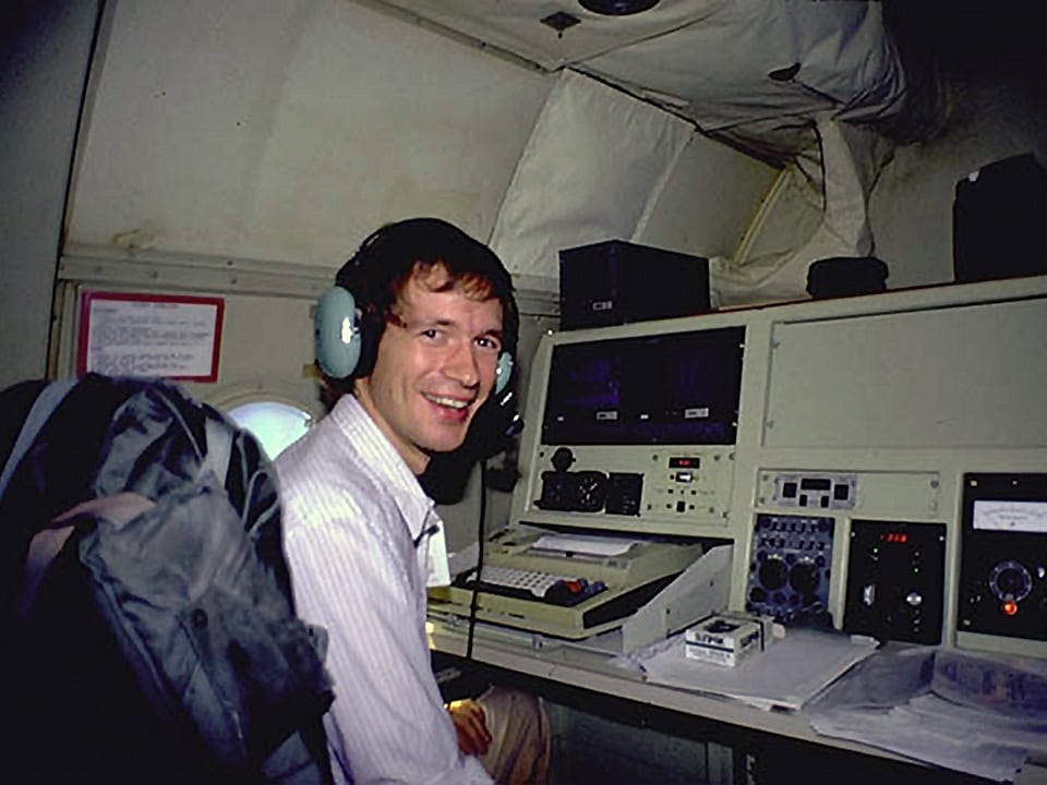 Jeff Masters seated at an electronic console with various buttons, switches, dials and an electric typewriter in a larger, non-commercial airplane next to a small window. He is looking at the camera, smiling and wearing a headset.