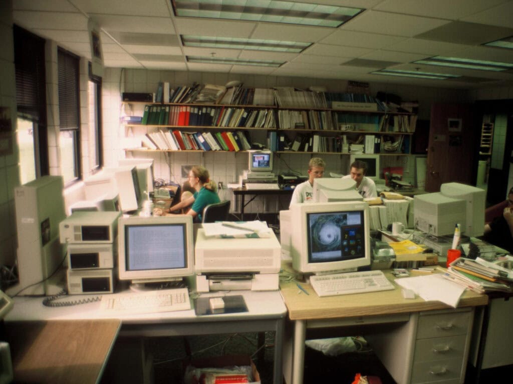 A room crowded with about eight 90's-era computers on desks and a few students sitting at them. A satellite image of a hurricane is on one of the monitors.