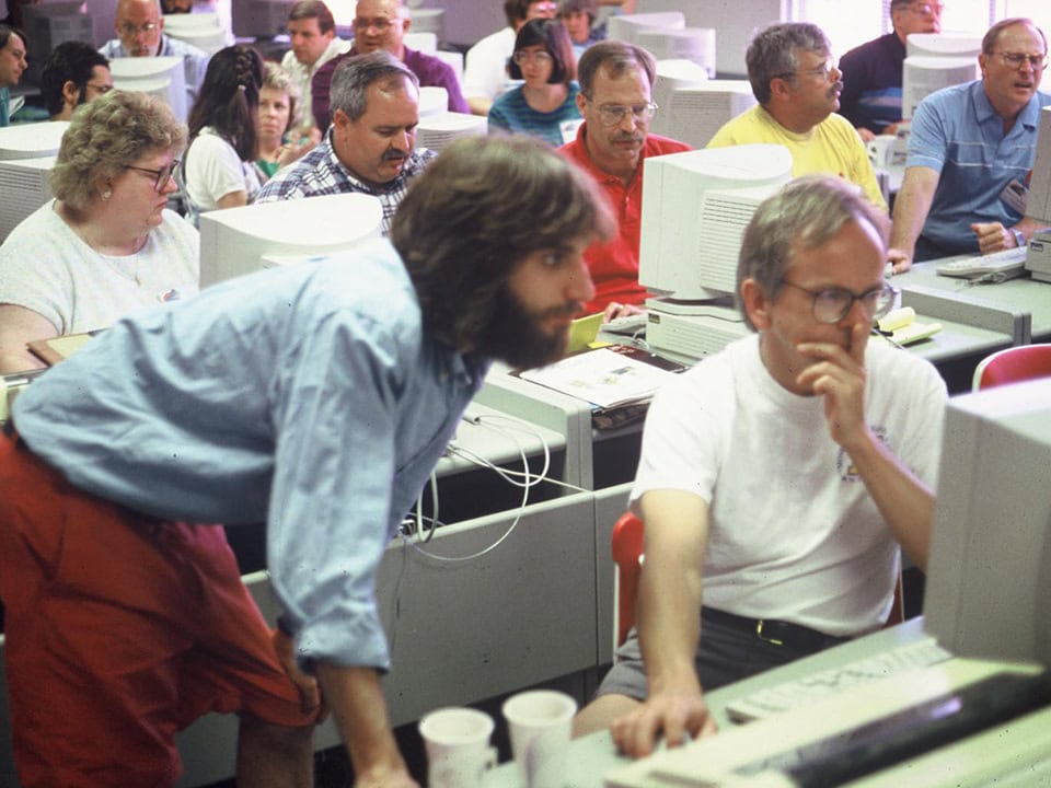Perry Samson and Jeff Ferguson looking intently at a 90s era computer screen while rows of adults at computers sit behind them.