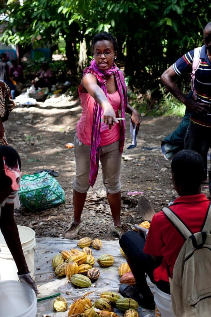 A woman points at cacao pods laid out on the ground.