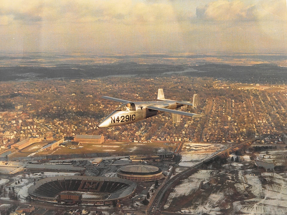 Scanned photograph of a small, un-painted aluminum plane with a propeller behind the tail flies over Michigan Stadium and Ann Arbor.
