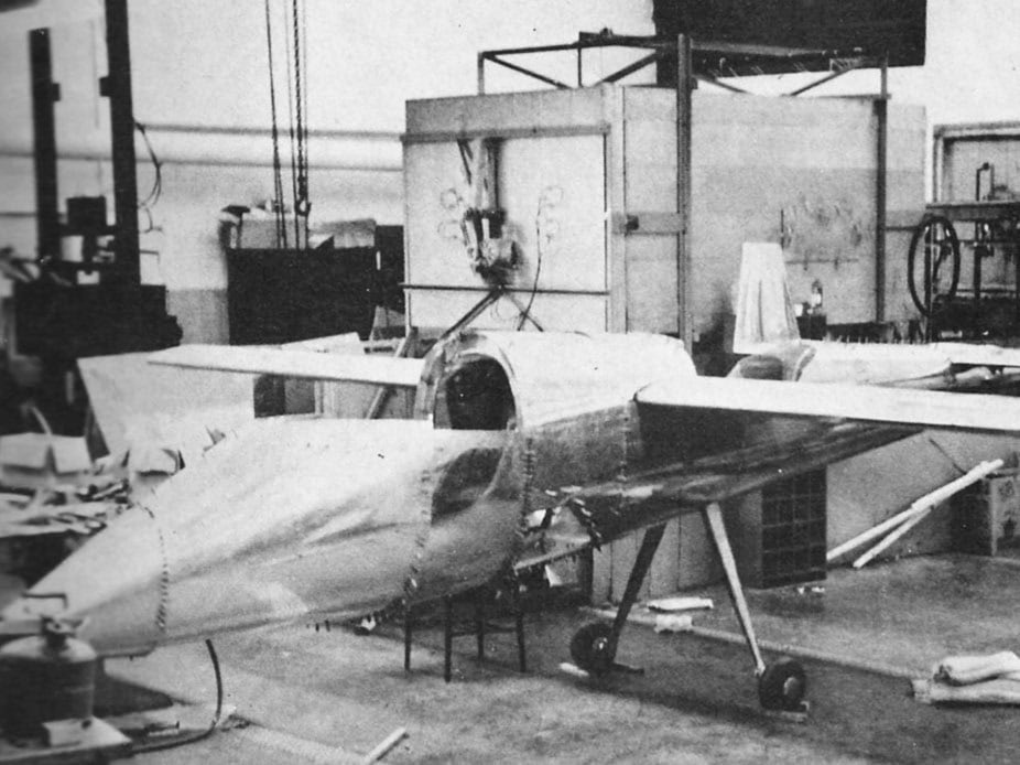 Black and white photo of the Teal partially constructed in lab that appears to barely have  enough space for the plane.