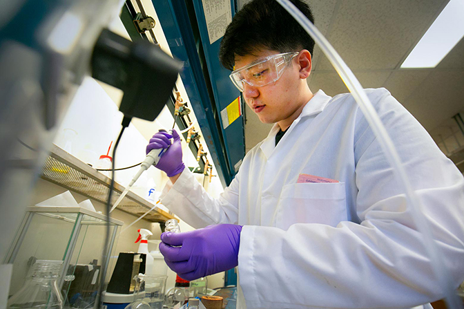 A man in a labcoat and safety goggles holding a micropipette.