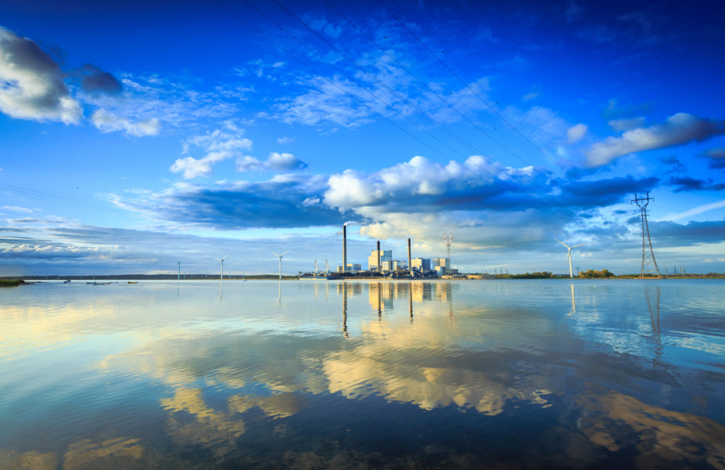 Buildings, wind turbines, and power lines in the distance reflected on a large body of water.