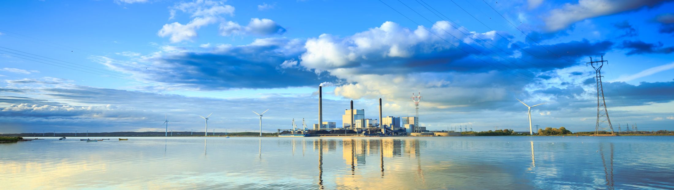 Buildings, wind turbines, and power lines in the distance reflected on a large body of water.