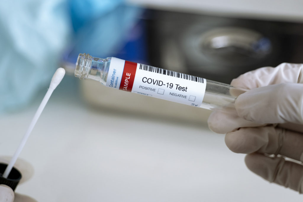 A person in gloves holds up a q-tip to a vial labeled "COVID-19 Test"