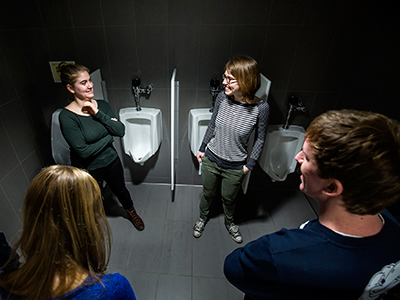 Four people standing by the urinals in a restroom.