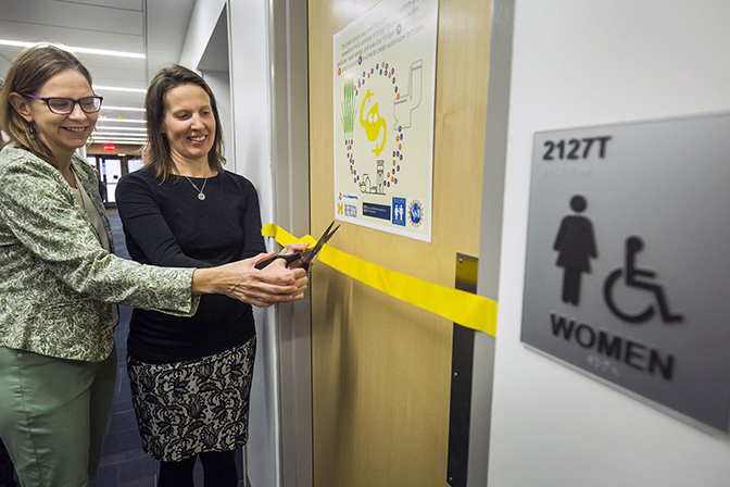 Two women cutting a yellow ribbon across a women's restroom door.