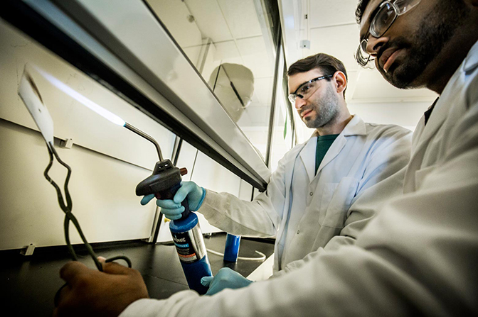 Two researchers, one holding a sheet in tongs, and the other holding a blowtorch towards it.