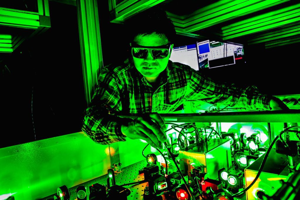 A man in safety goggles adjusts wiring in a dark room with green lasers.