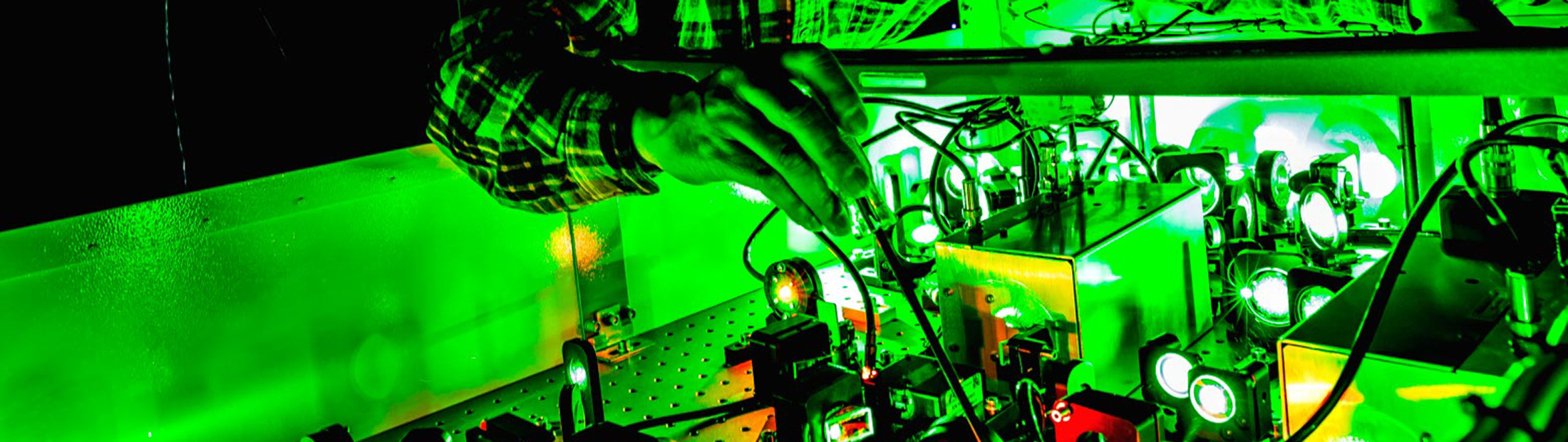 A man in safety goggles adjusts wiring in a dark room with green lasers.