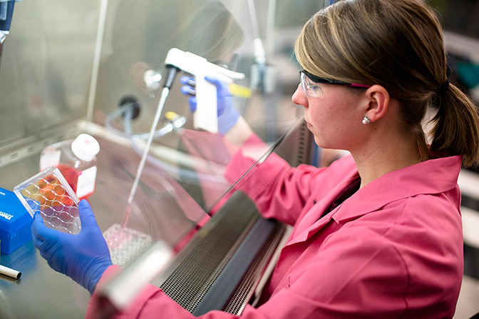 A woman in a labcoat and goggles holding lab equipment.