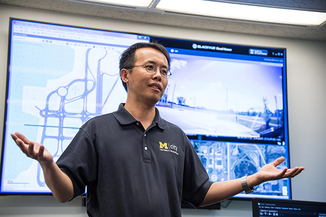 A man gesturing in front of a screen displaying roads and diagrams.