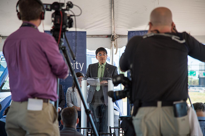 People with cameras face a man speaking at a podium.