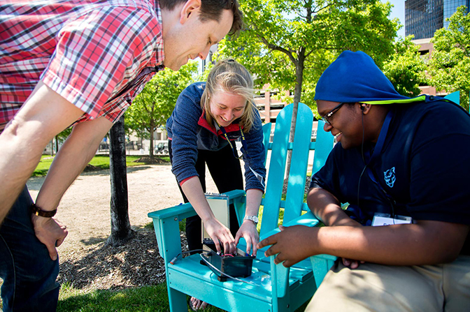 Three people gather around a device places on a blue chair.