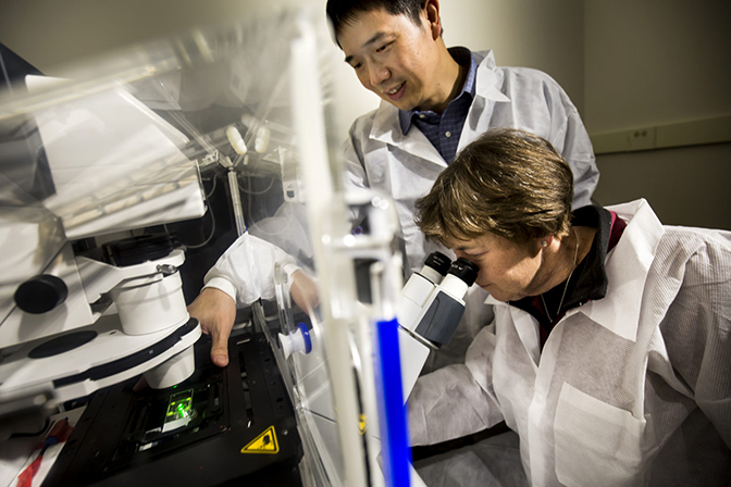 A woman in a labcoat looking through a microscope while a man in a labcoat stands beside her.