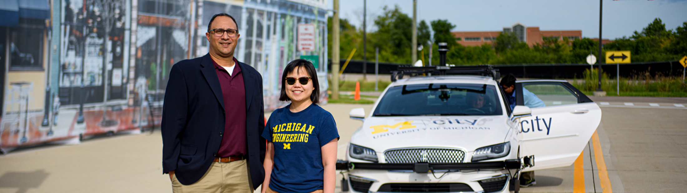 Two people standing on a road with a car labelled "Mcity" behind them.
