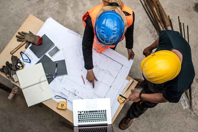 Two people in hardhats at a workbench with blueprints and a laptop.