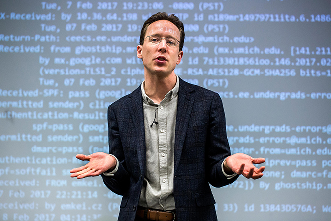 A man speaking in front of a screen with code on it.