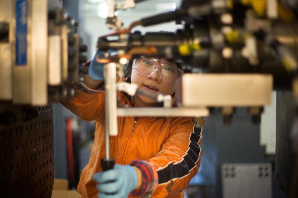 A woman in safety goggles holding a screwdriver up to a machine.