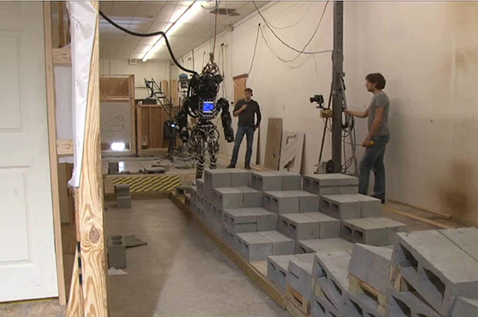 A human-sized bipedal robot moves through a room full of cinderblocks.