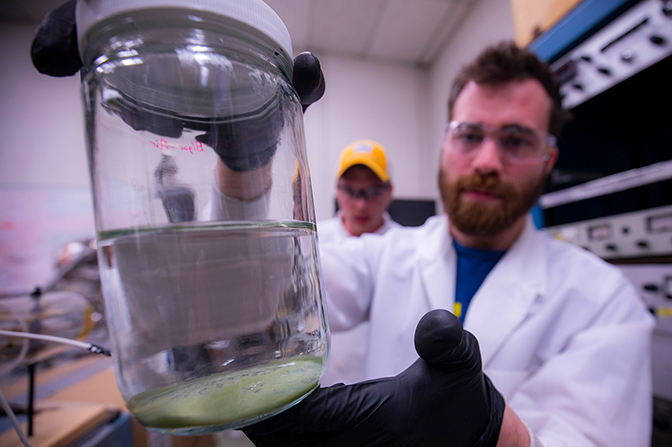 A man in a labcoat and goggles holding a beaker of clear liquid.