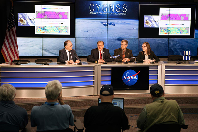 Four people holding a press conference with reporters seated in the audience.