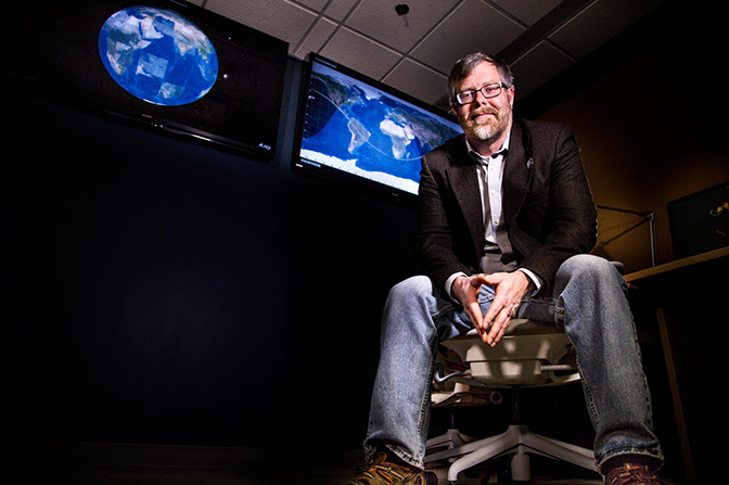 A man leaning forward in a chair facing the camera, with screens displaying a world map behind him.