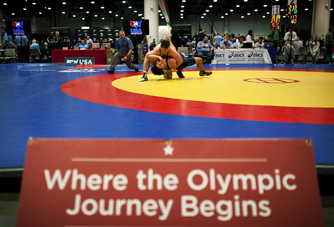 Two people wrestling in a ring with a sign reading "Where the Olympic Journey Begins" visible in the foreground.