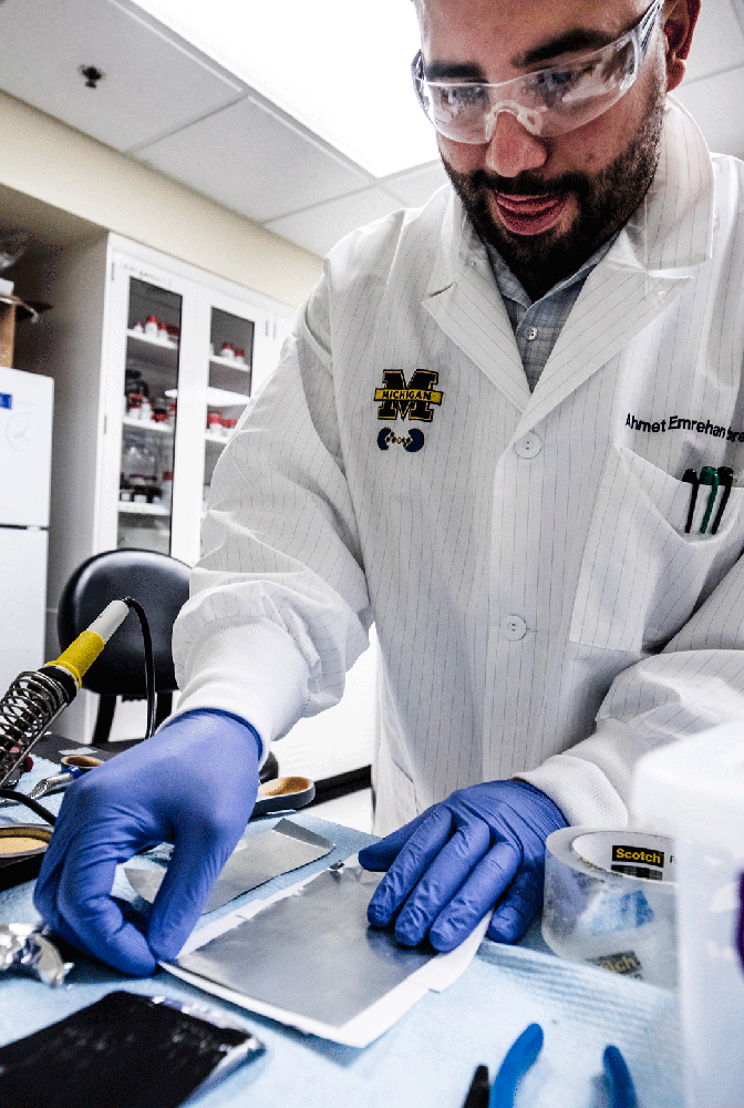 A man in a labcoat and goggles handling a thin sheet of metal.