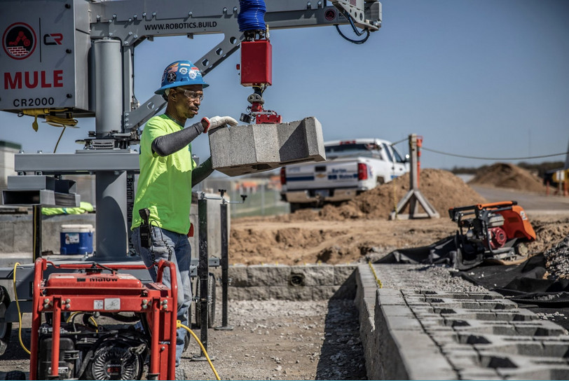 With the help of MULE, a simple robot, a construction worker places a concrete block in a retaining wall.