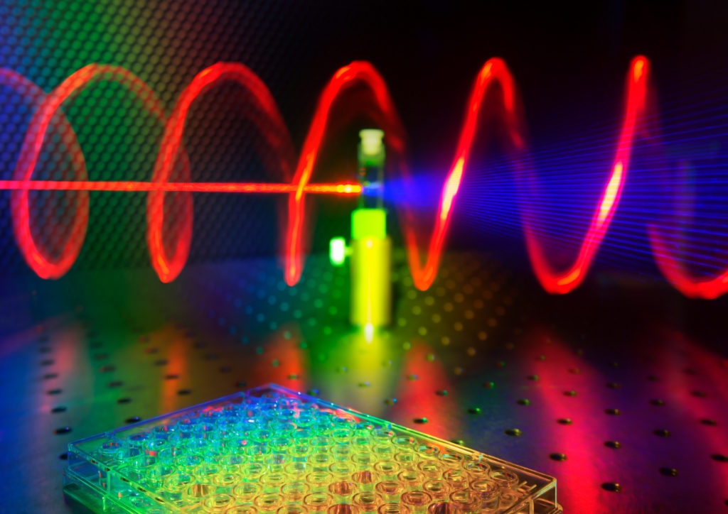 The illustration shows a microplate well in the foreground, while in the background a tested sample receives red laser light and releases twisted blue light. Credit: Ventsislav Valev, Kylian Valev and Lukas Ohnoutek, University of Bath.