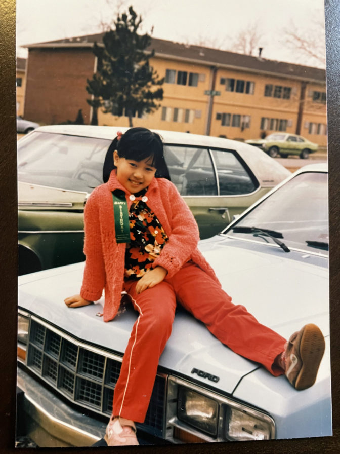 Linda Zhang as a young child wearing an orange jumpsuit sitting on the hood of a car