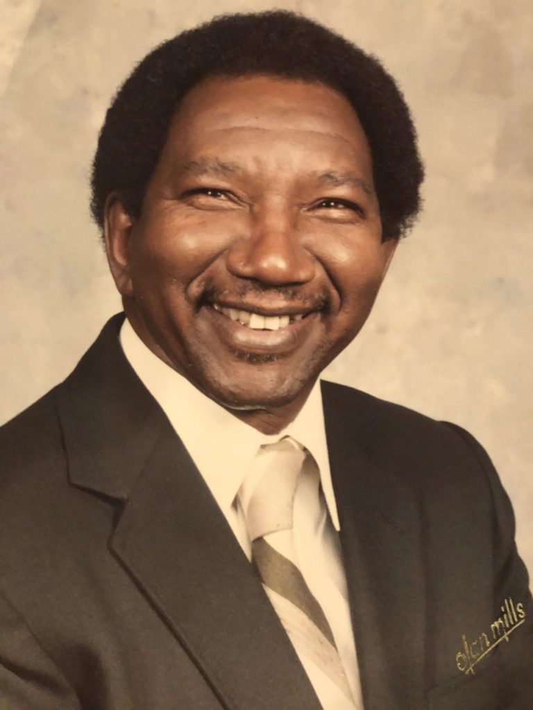 An elderly Black man smiling for a photo wearing a black suit with a striped tie and a white button up shirt