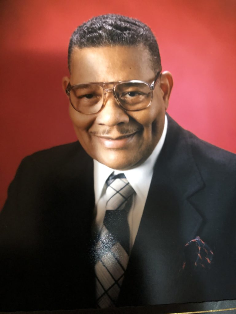 An elderly Black man smiling for a photo wearing a in a black suit and a plaid tie against a red backdrop