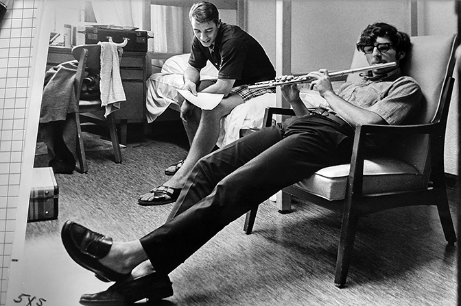 A student plays a flute in his dorm room while another student looks at a sheet of paper while sitting on the bed.