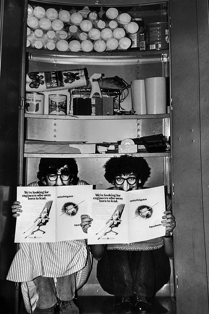 Two men crouching on the bottom of a shelf displaying a Michigan Techinic pamphlet.