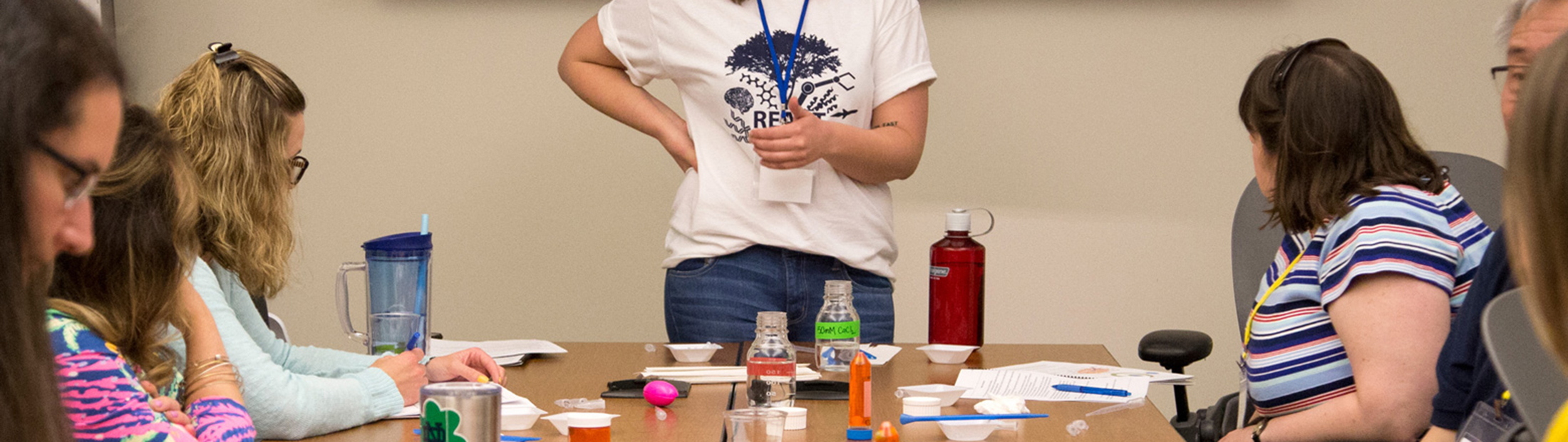 A woman standing at the head of a table filled with people doing an activity with pens and paper.