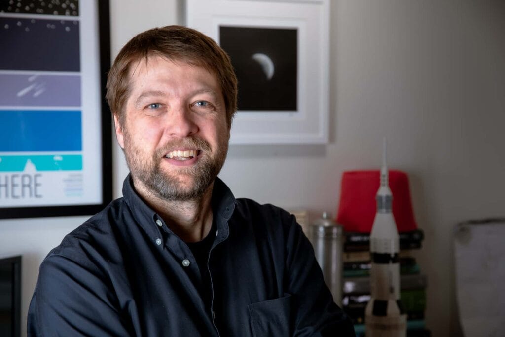 A man with short hair and beard smiles, wearing a navy dress shirt in his neat office.
