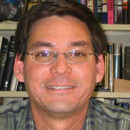 Portrait of Mark Moldwin - he's wearing a button up shirt, wears glasses and has short hair. Behind him is a bookshelf.
