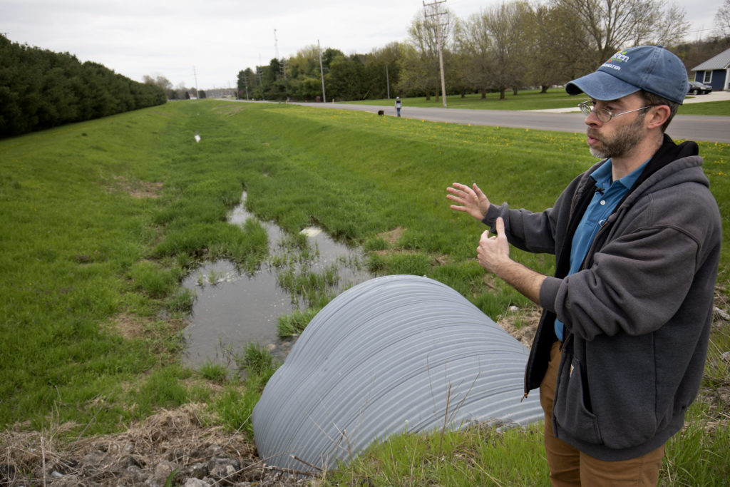 A man stands over a drainage ditch wearing a blue hat and grey jacket. the drainage field has a small amount of water in it.