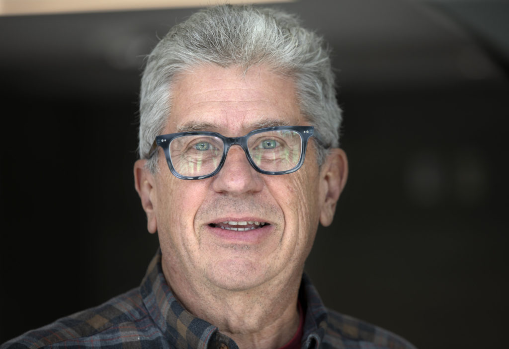 Richard Rood smiling, wearing thick black glasses, a plaid shirt. He has short, neat grey and black hair and stands in a dark office.