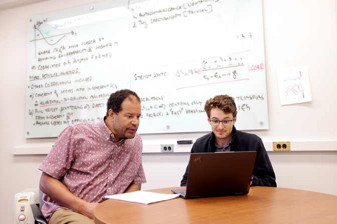 Two men sit at a table reviewing data on a computer, behind them is a white board covered in equations.