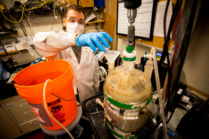 A masked and goggled researcher pours an orange bucket and combines materials.