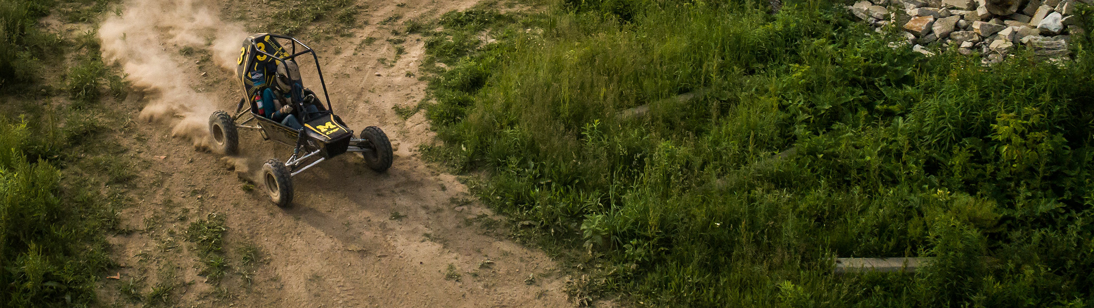 An off-road Baja racing vehicle kicks up dust on a dirt path next to some long, green grass.