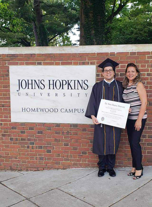 Díaz is wearing a graduation cap and gown and holding a large diploma while standing between his mother and a sign that reads, "Johns Hopkins University Homewood Campus".
