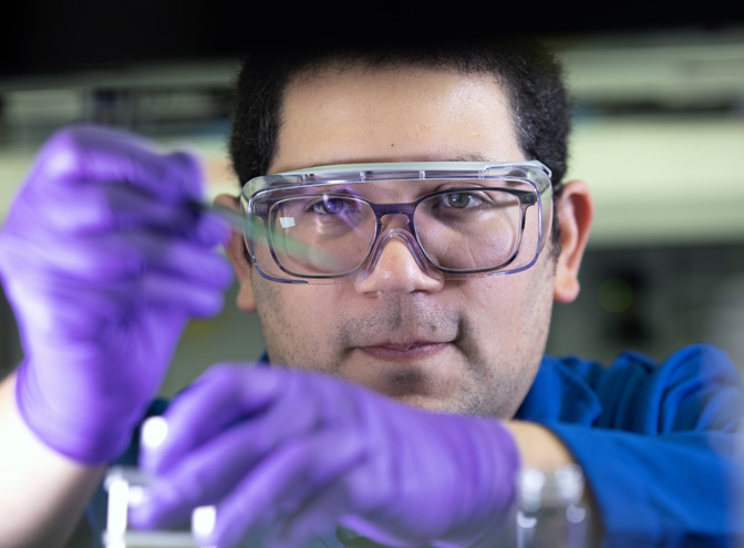 Díaz is wearing safety glasses and using his gloved hand to hold up an out-of-focus object.