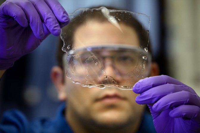 Díaz, wearing safety glasses, looks through a roughly square, transparent material while wearing purple gloves.